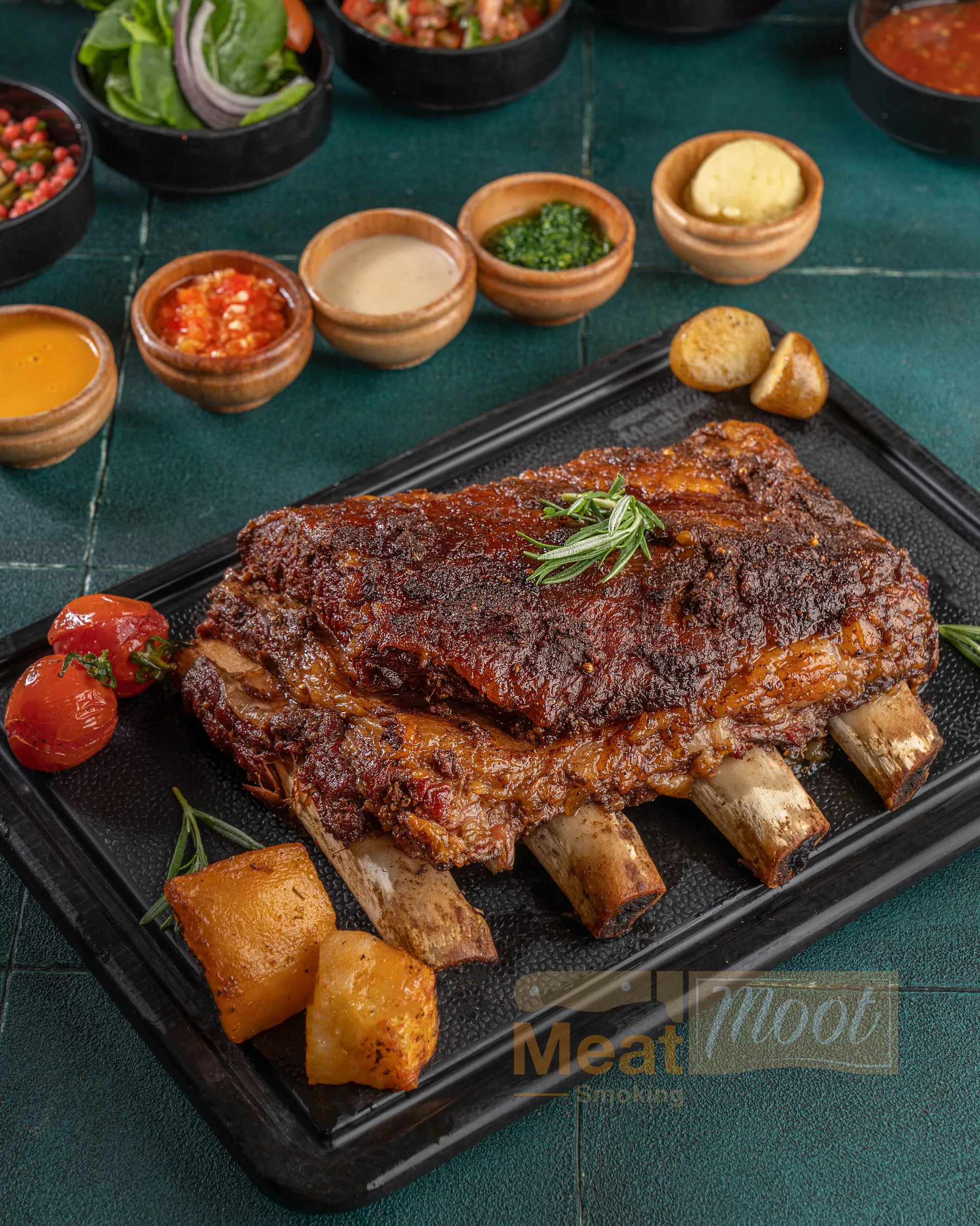 A rack of smoked halal beef ribs on a black platter with rosemary, roasted potatoes, cherry tomatoes and house sauces