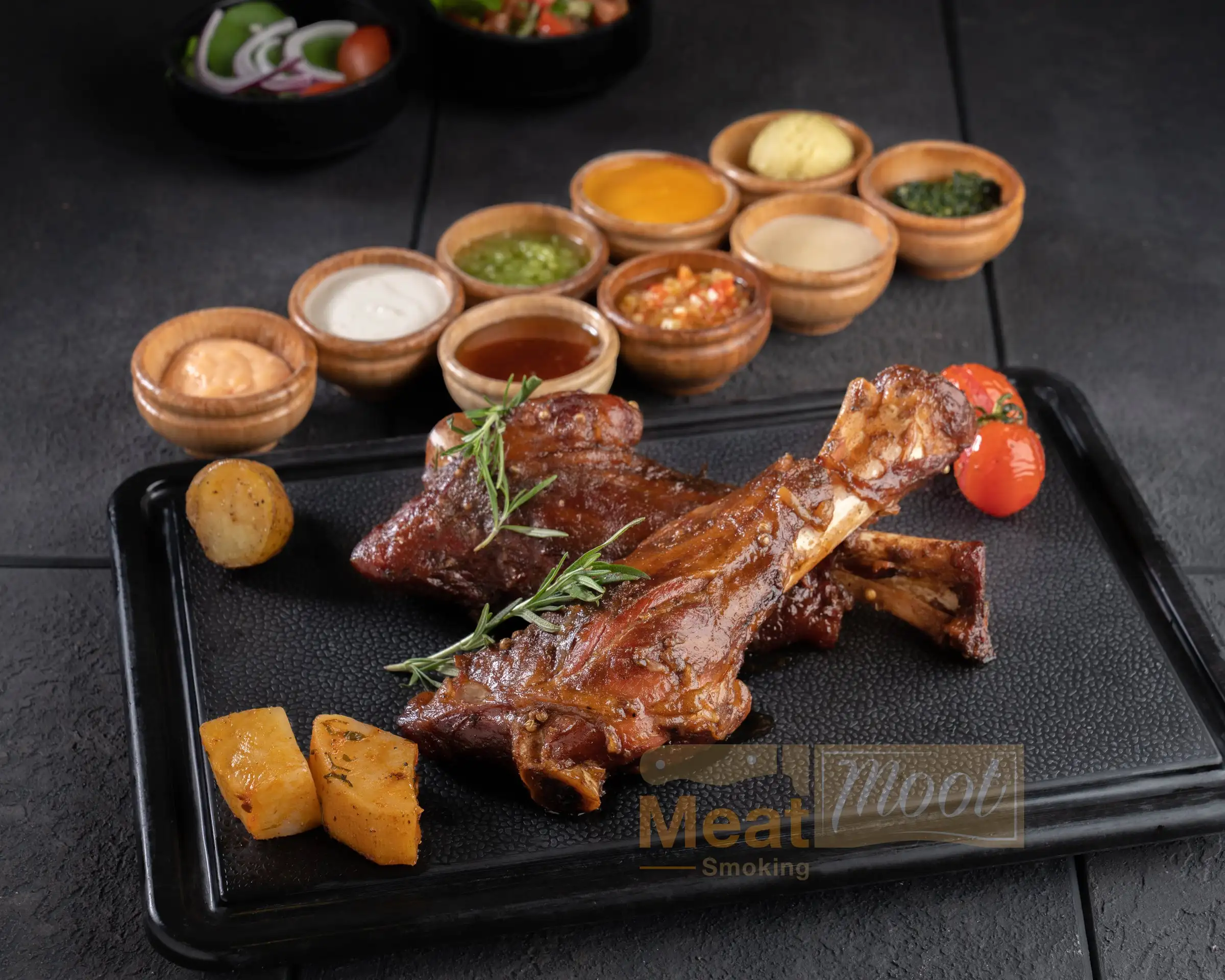 Smoked lamb shanks on a black platter with rosemary, roasted potatoes, cherry tomatoes and house sauces at Meat Moot Seattle